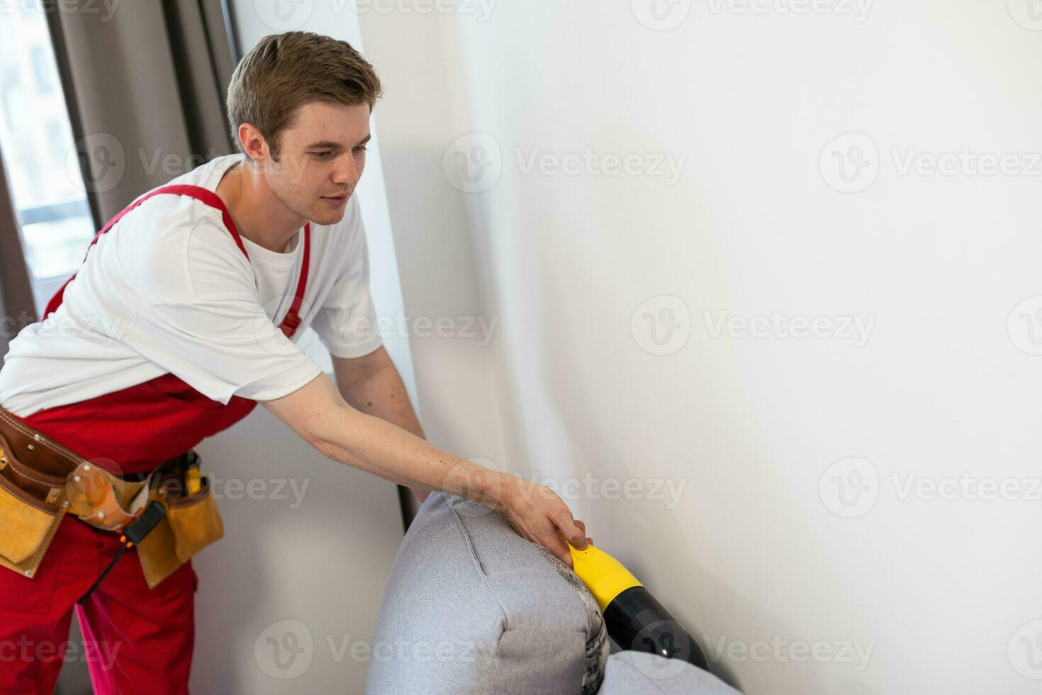 achterzijde visie van jong mannetje arbeider schoonmaak sofa met vacuüm schoonmaakster foto