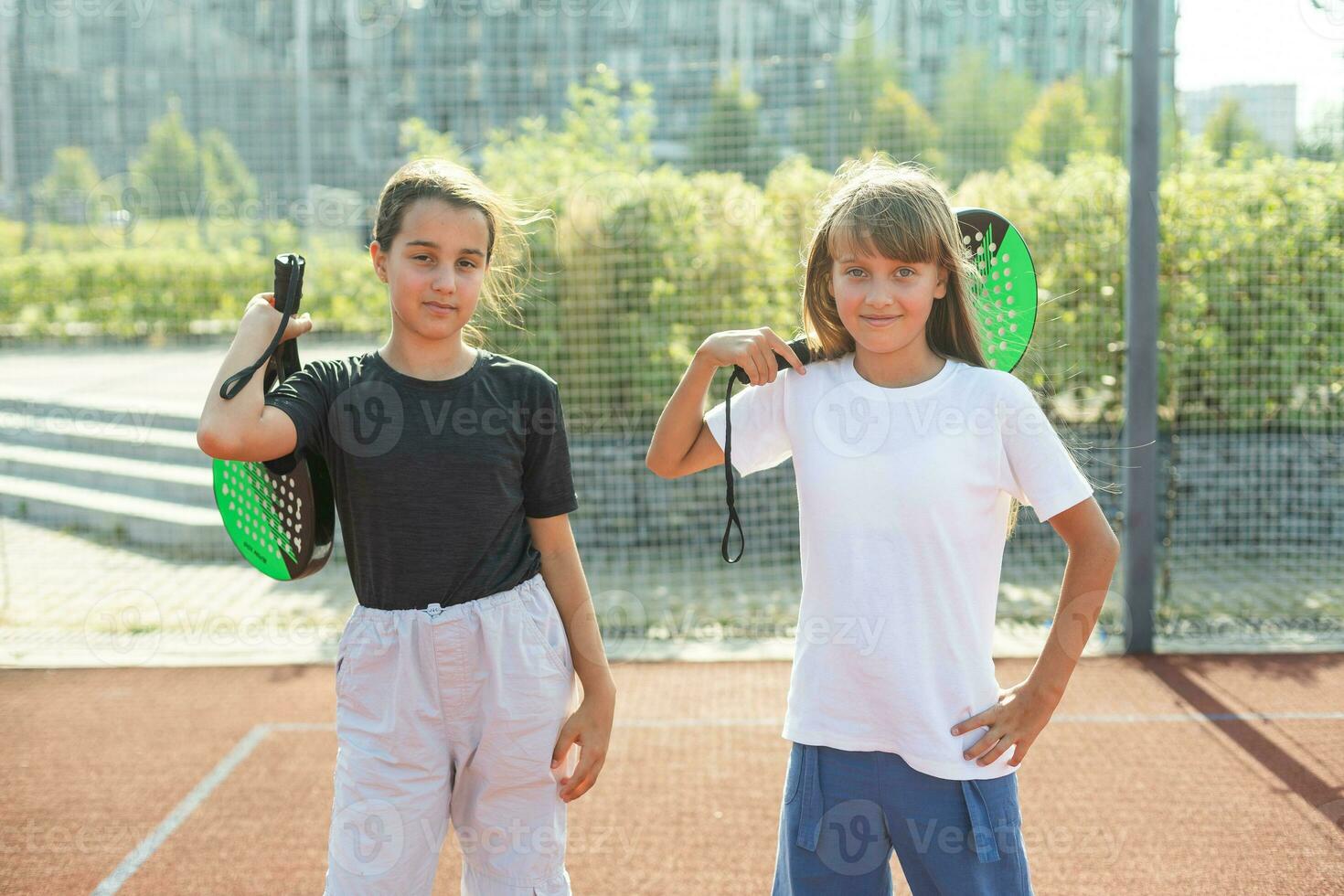 kinderen en sport- concept. portret van glimlachen meisjes poseren buitenshuis Aan padel rechtbank met rackets en tennis ballen foto