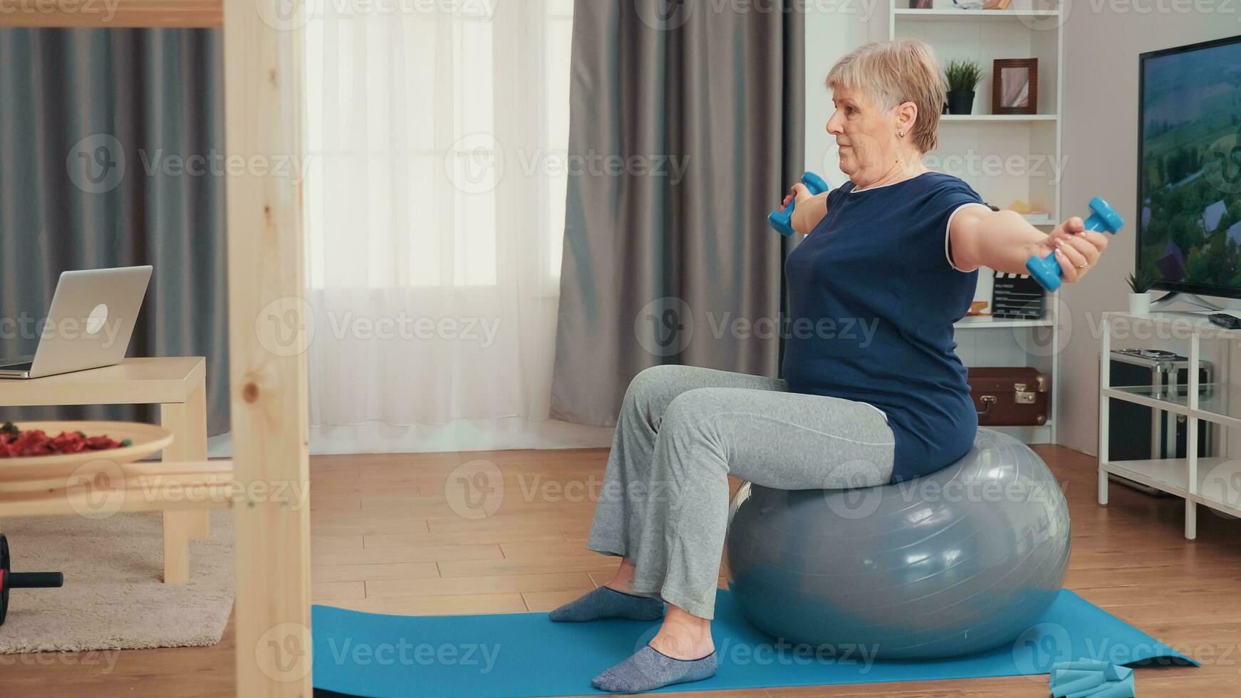 grootmoeder hijs- gewichten zittend Aan balans bal in leven kamer. oud persoon opleiding Bij huis sport gezond levensstijl, ouderen geschiktheid oefening training in appartement, werkzaamheid en gezondheidszorg foto