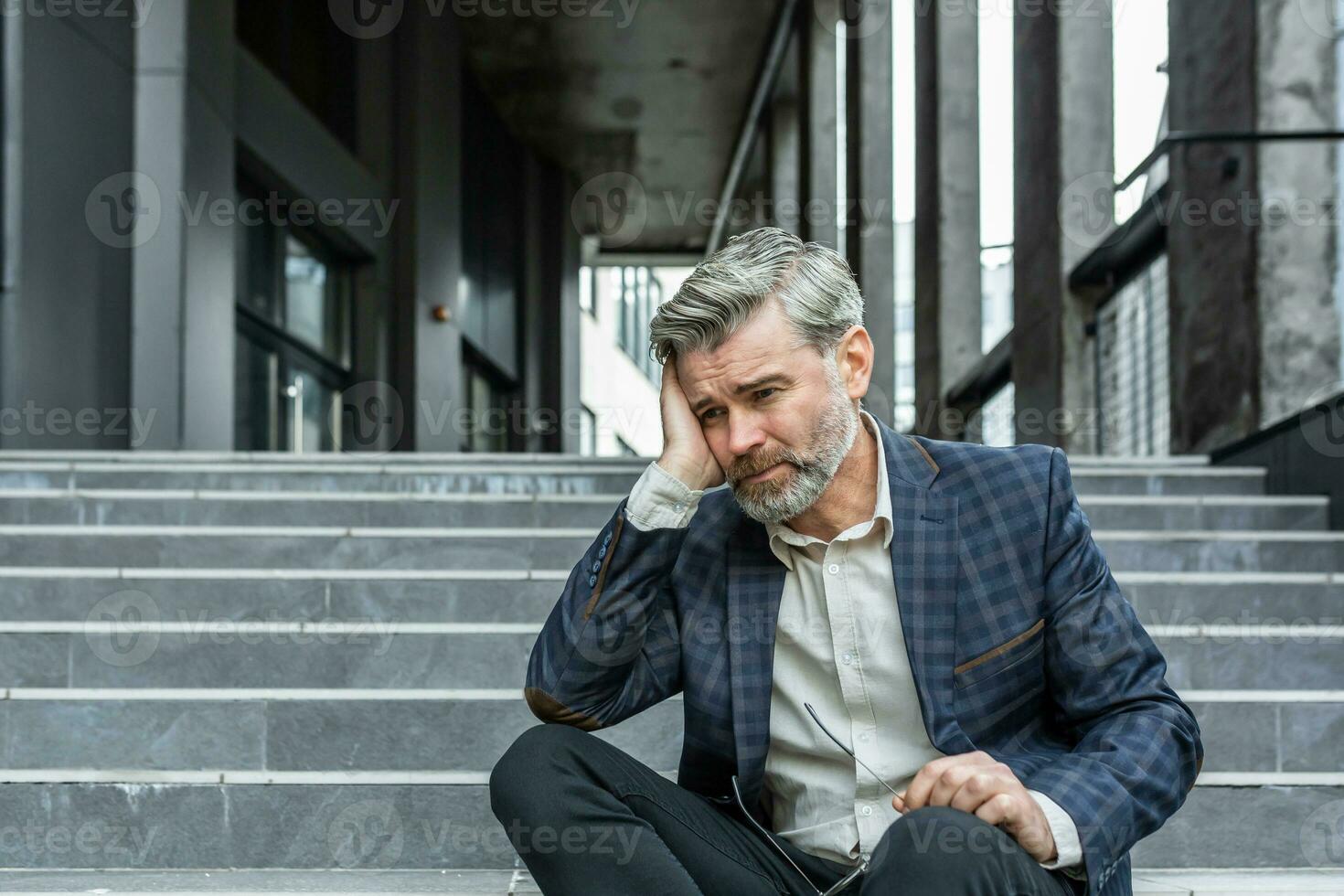 grijs haren zakenman zittend verdrietig Aan trap buiten kantoor gebouw, depressief baas in bedrijf pak van streek en teleurgesteld met prestatie resultaten. foto