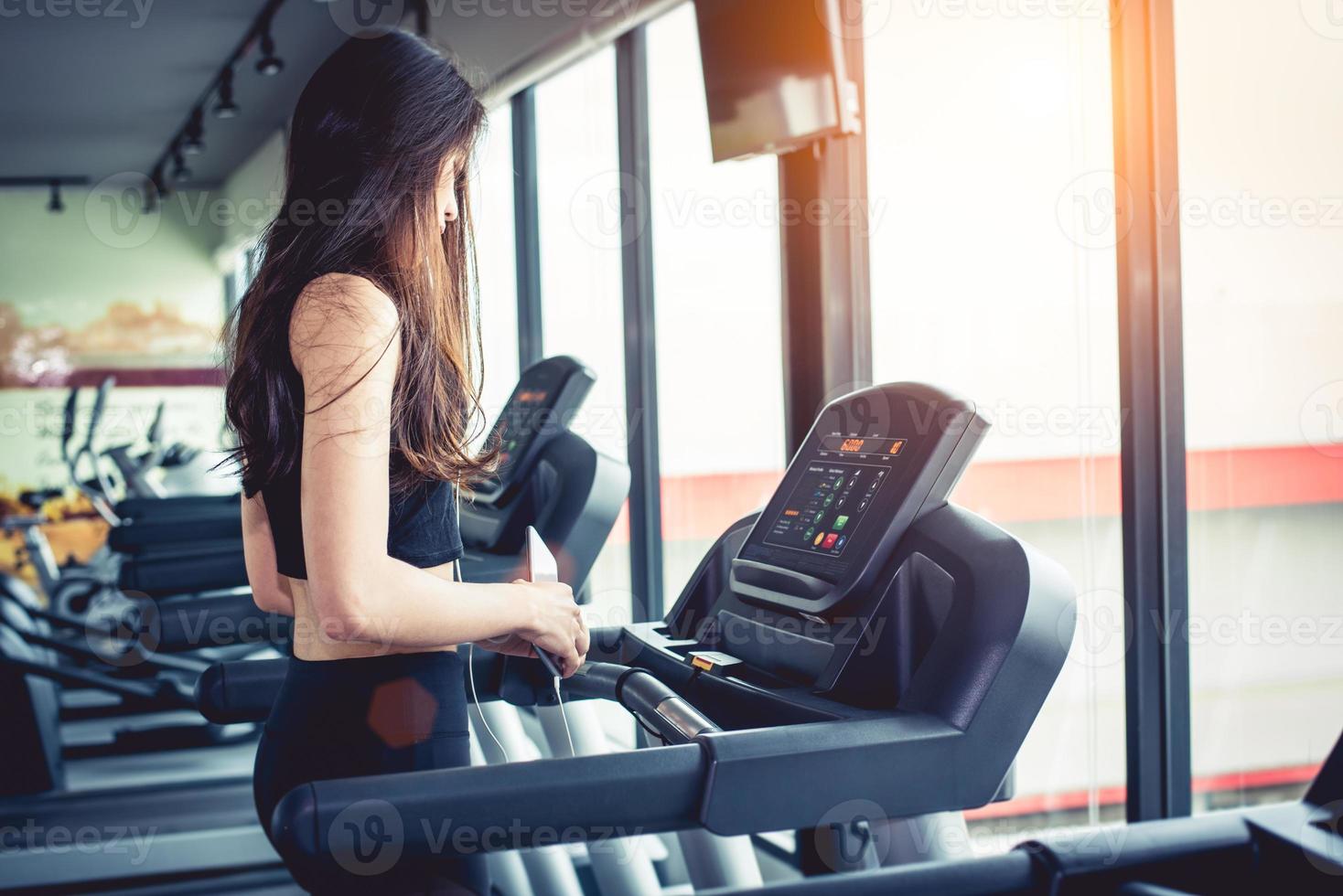 aziatische vrouw die slimme telefoon gebruikt tijdens training of krachttraining bij fitness gym op loopband. ontspannen en technologie concept. sport oefening en gezondheidszorg thema. gelukkig en comfortabel humeur foto