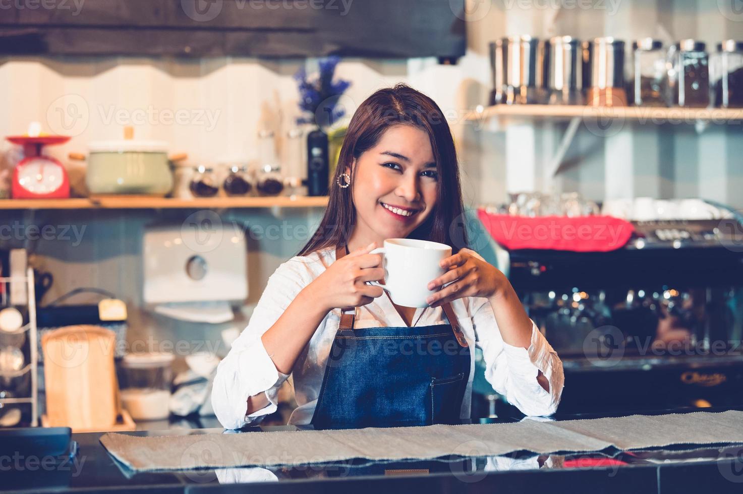 Aziatische vrouwelijke barista kopje koffie maken. jonge vrouw die een witte koffiekop vasthoudt terwijl ze achter de cafébar op de achtergrond van het restaurant staat. mensenlevensstijlen en bedrijfsbezettingsconcept foto