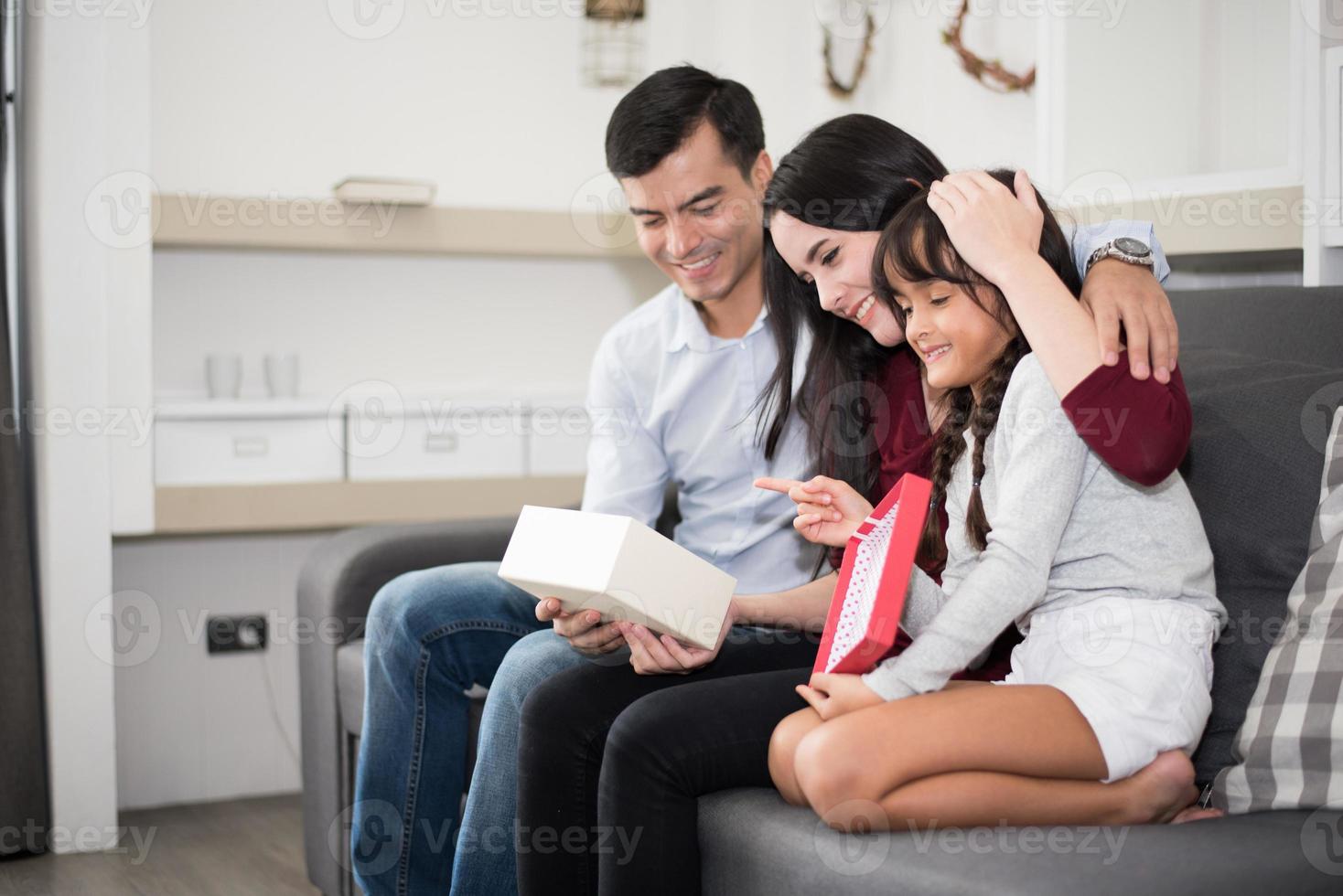 vader en moeder verrassen hun dochter met een cadeau of nieuw speeltje. ouders en kinderen zijn gelukkig samen in huis op de bank. familie en geluk concept foto