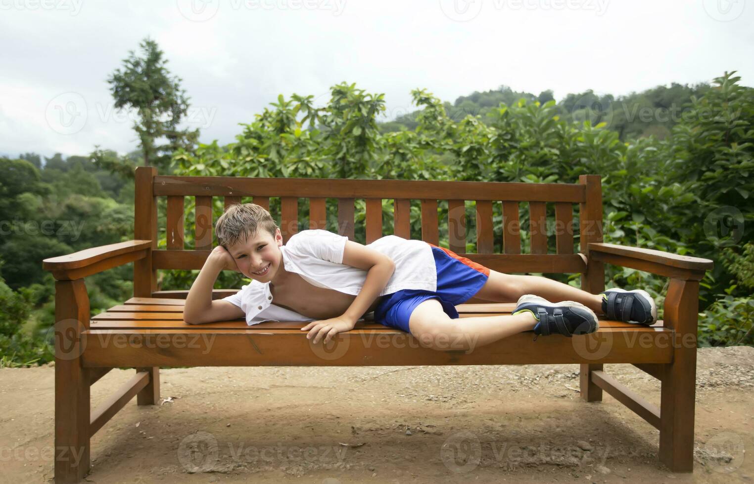 een gelukkig kind in de zomer warmte leugens Aan een houten bank tegen de backdrop van tropisch planten. de jongen is Aan vakantie in de zomer. foto