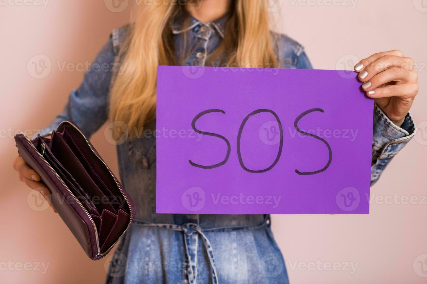 vrouw Holding leeg portemonnee en Sos woord geschreven Aan papier terwijl staand in voorkant van muur. foto