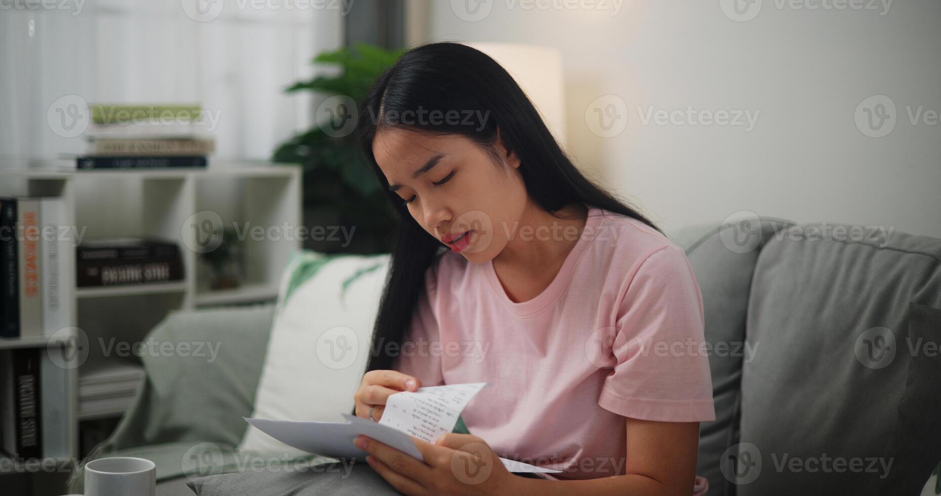 portret van jong Aziatisch vrouw gefocust in berekenen begroting zittend Aan sofa Bij huis Holding papier belastingen rekeningen en lezing financieel informatie, thuis financiën foto