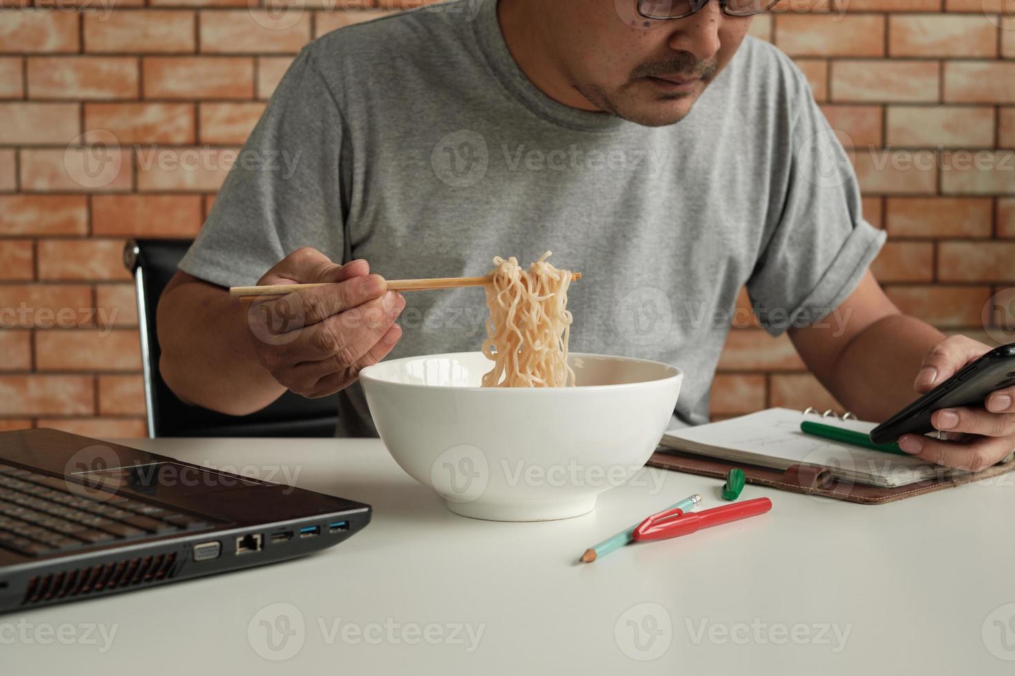 mannelijke werknemer druk aan het werk met een laptop en mobiele telefoon, gebruik eetstokjes om instant noedels te eten tijdens kantoorlunch, omdat het snel, smakelijk en goedkoop is. na verloop van tijd Aziatisch fastfood, ongezonde levensstijl. foto