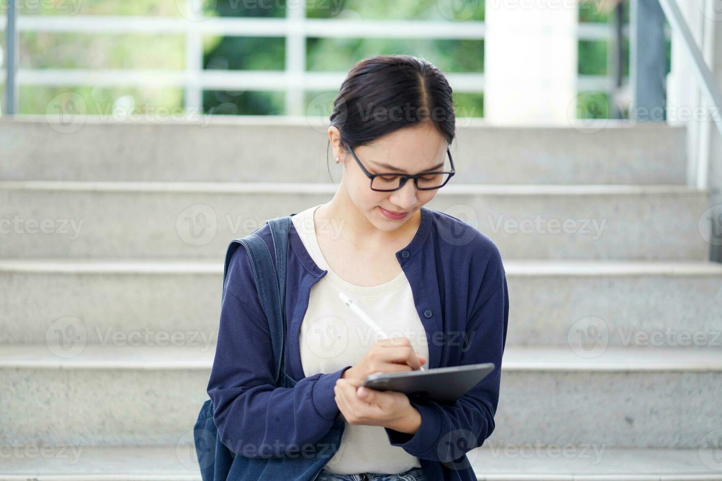 detailopname Aziatisch jong vrouw leerling in gewoontjes kleding lezing en maken een kort Notitie Aan tablet voor de tentamen Aan de trap van de school- gebouw en wazig achtergrond. Aziatisch school- concept. foto