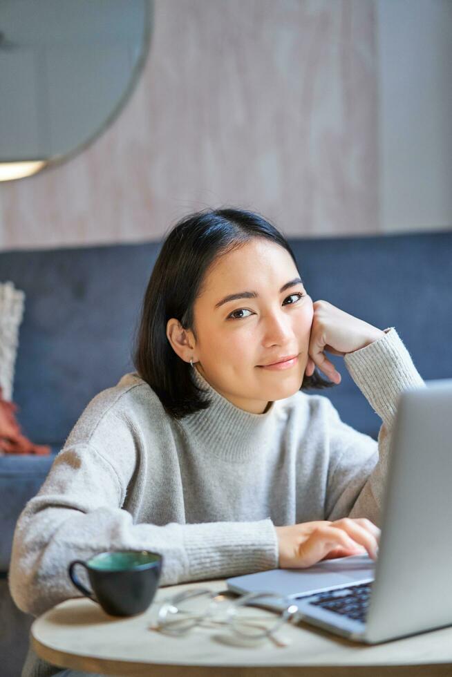 verticaal schot van jong Aziatisch vrouw op zoek moe, glimlachen met uitgeput uitdrukking, werken van huis Aan haar laptop foto