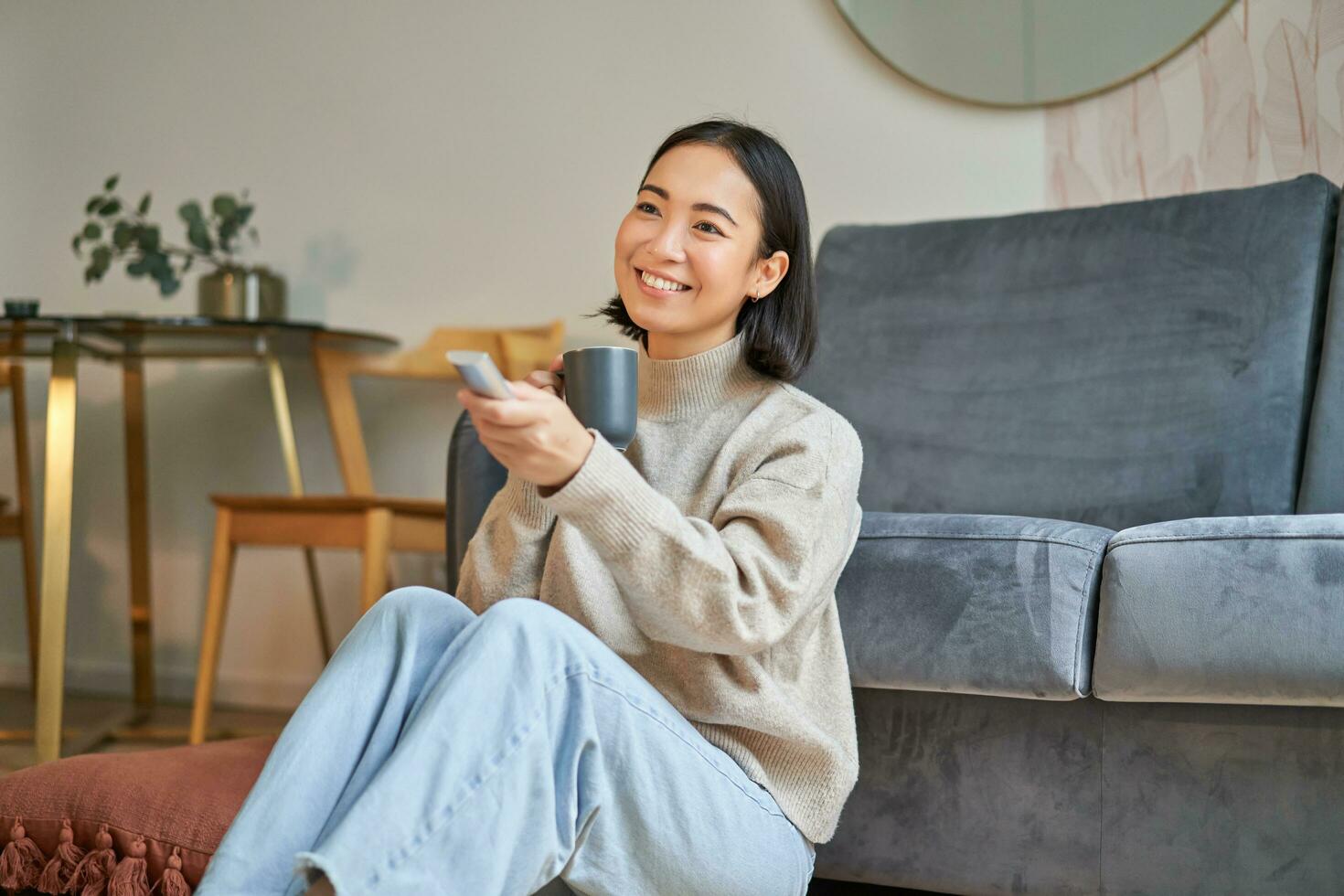 portret van glimlachen jong vrouw resting Bij huis in de buurt TV, aan het kijken televisie zittend Aan verdieping en drinken koffie van kop foto