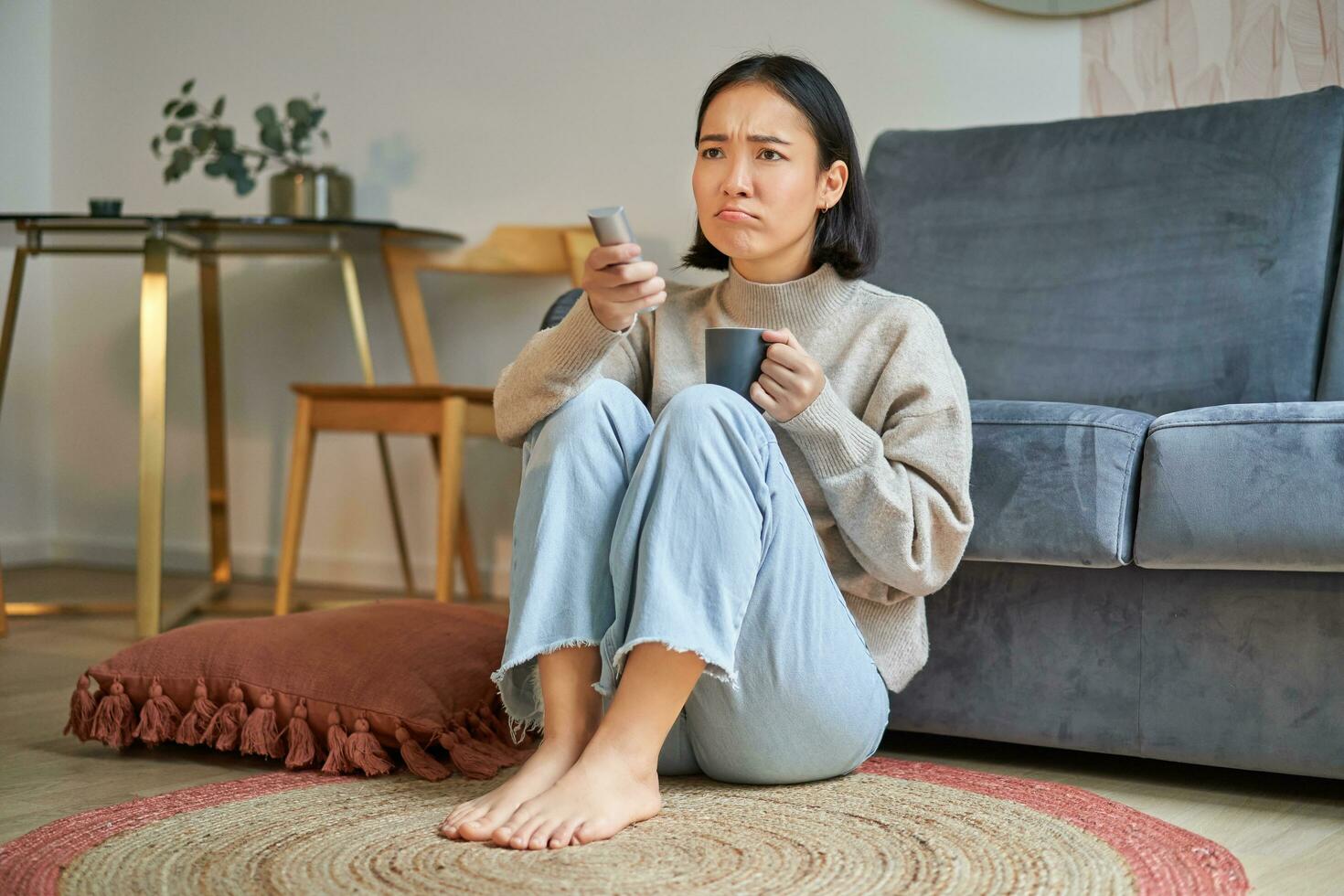 portret van jong vrouw met op afstand, aan het kijken TV, schakelen kanalen Aan televisie, zittend Aan verdieping in de buurt sofa en ontspannende foto