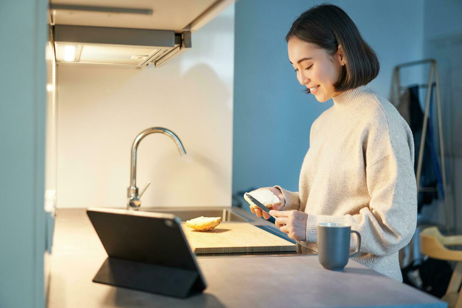 portret van jong Koreaans vrouw, maken geroosterd brood en aan het kijken video Aan digitaal tablet, op zoek Bij scherm foto