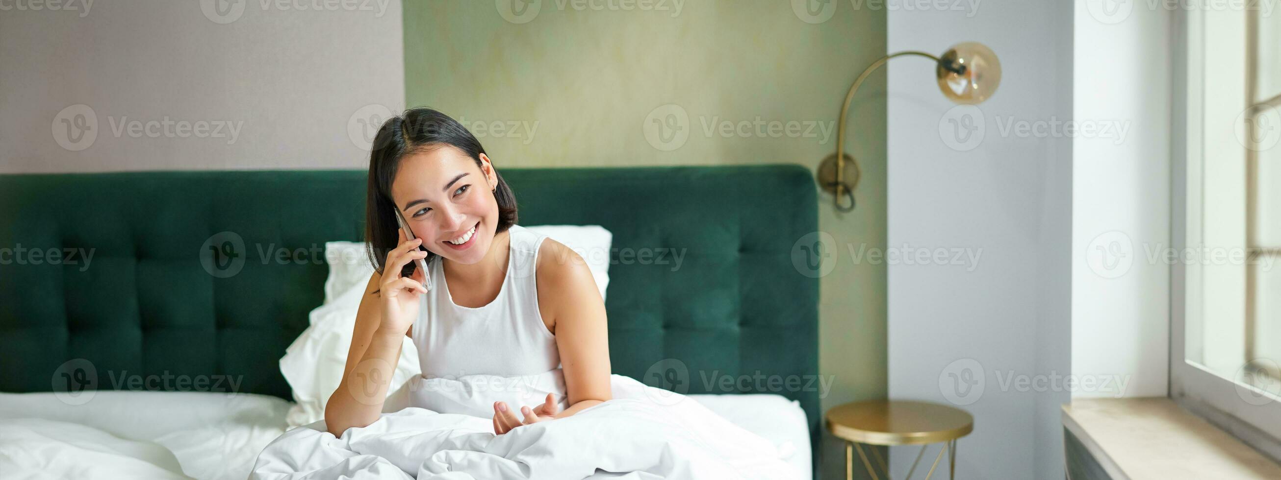 glimlachen Koreaans meisje praat Aan mobiel telefoon en aan het liegen in bed. schattig vrouw antwoorden telefoon telefoongesprek, houdt smartphone, ontspannende in haar bed foto