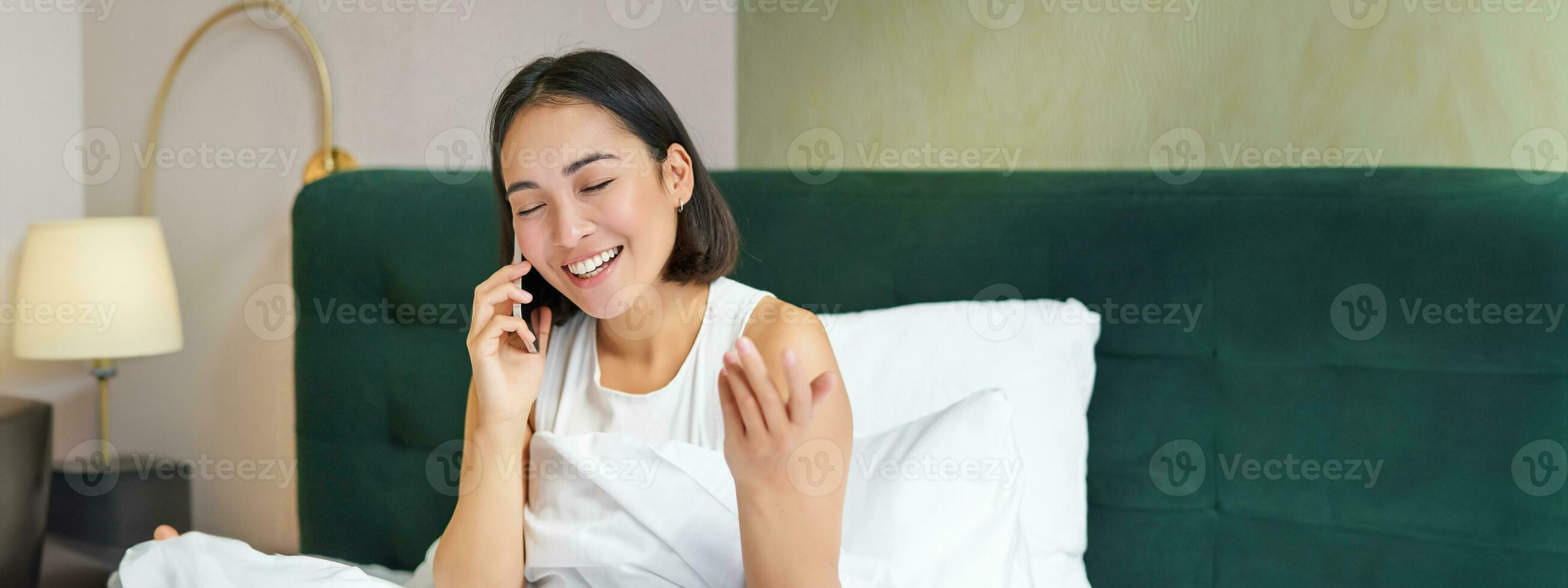 dichtbij omhoog portret van schattig Aziatisch meisje in bed, pratend Aan mobiel telefoon met gelukkig glimlachen gezicht. vrouw ontwaken omhoog en maken een telefoon telefoontje foto