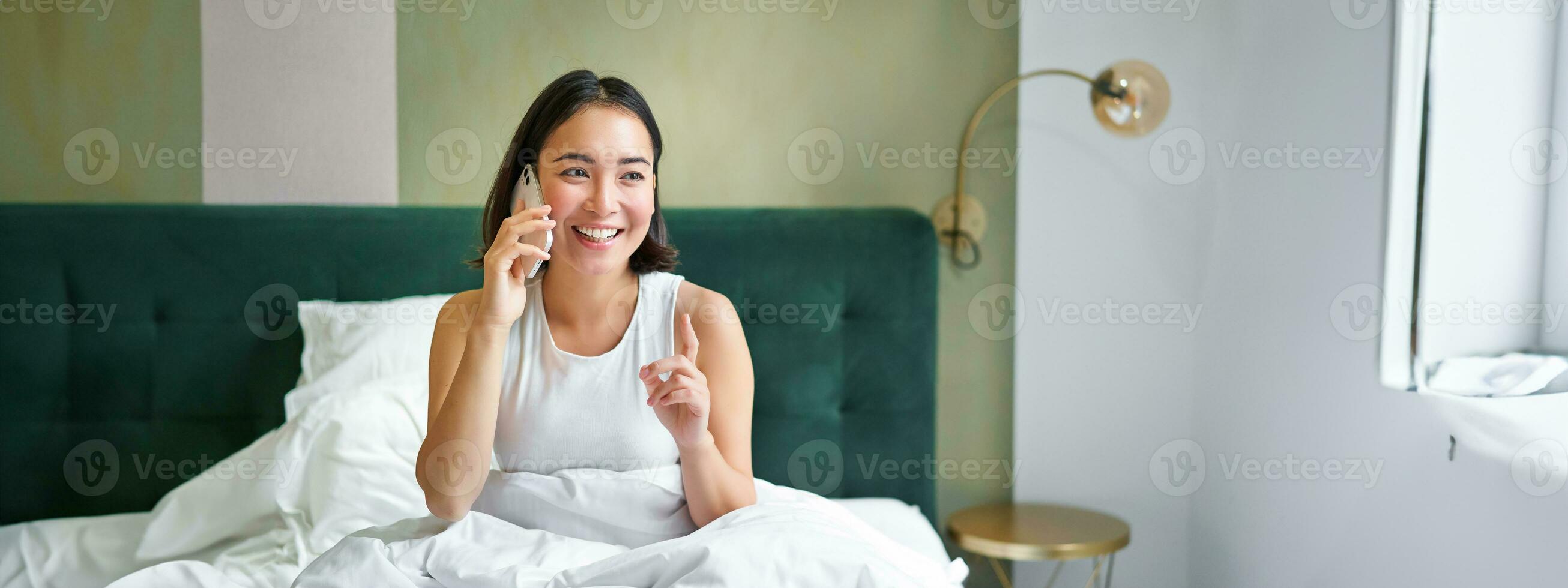 mooi glimlachen Aziatisch meisje pratend Aan mobiel telefoon, aan het liegen in bed met verheugd gelukkig gezicht, sprekend naar iemand Aan telefoon foto