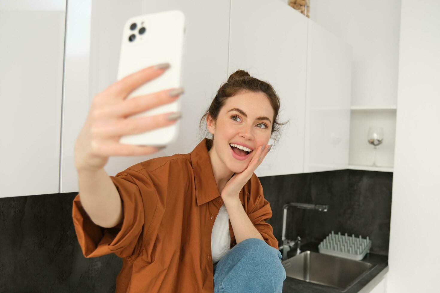 jong gelukkig vrouw nemen selfies Aan haar nieuw smartphone, poseren voor foto's met lachend, schattig gezicht uitdrukking, zittend in de keuken foto