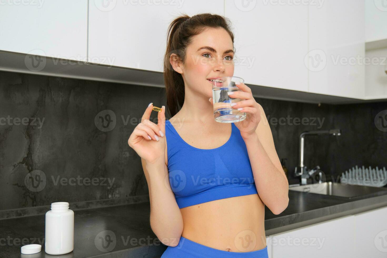 portret van mooi, brunette vrouw, vervelend geschiktheid actieve kleding, drinken water met dieet supplement, nemen vitamine d bloemknoppen voor gezond, sterk lichaam, staand in keuken foto