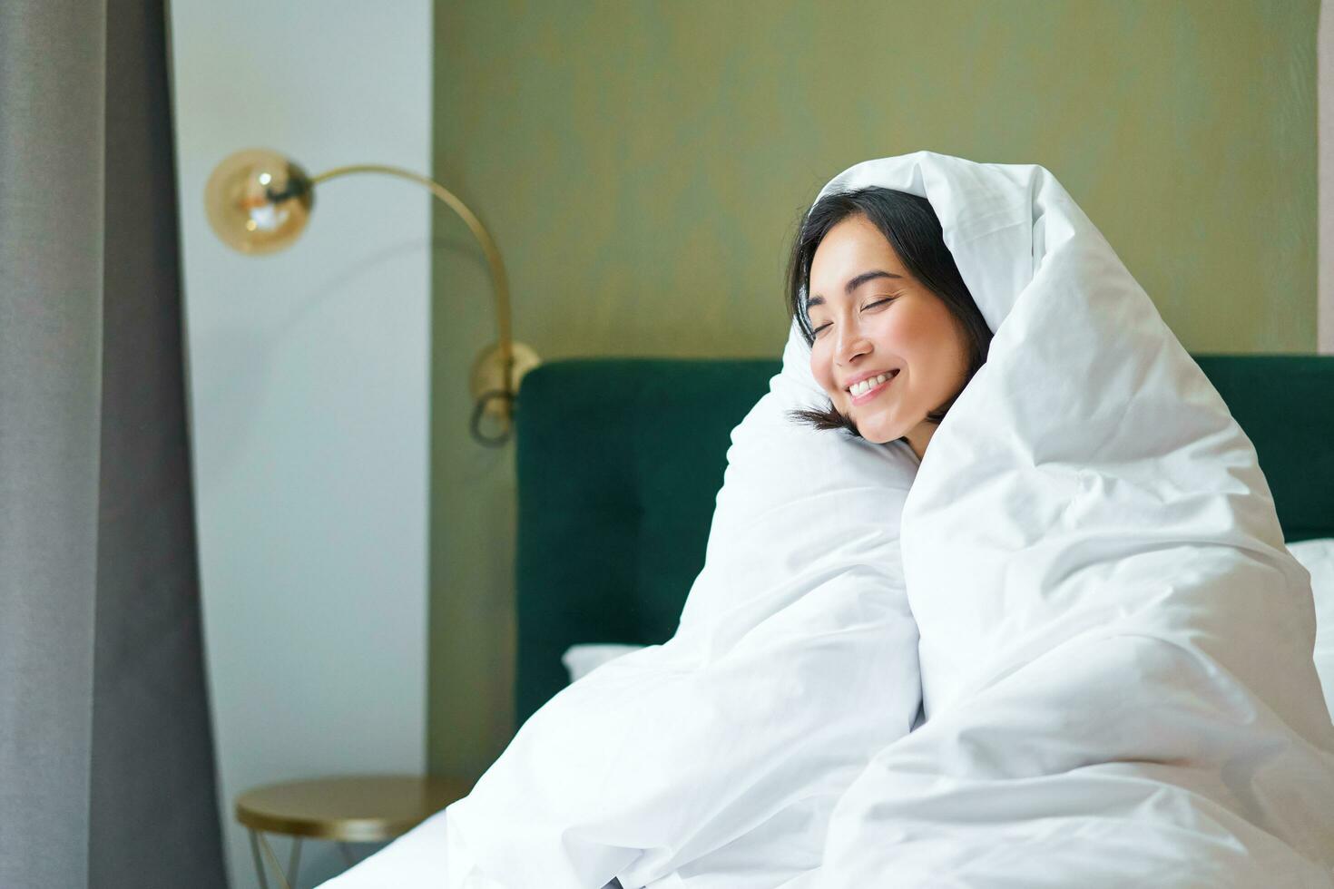 knus ochtenden. gelukkig Aziatisch meisje voelt warm in haar bed, covers haarzelf met knus dekbed in haar slaapkamer, een comfortabel blijven in hotel kamer foto