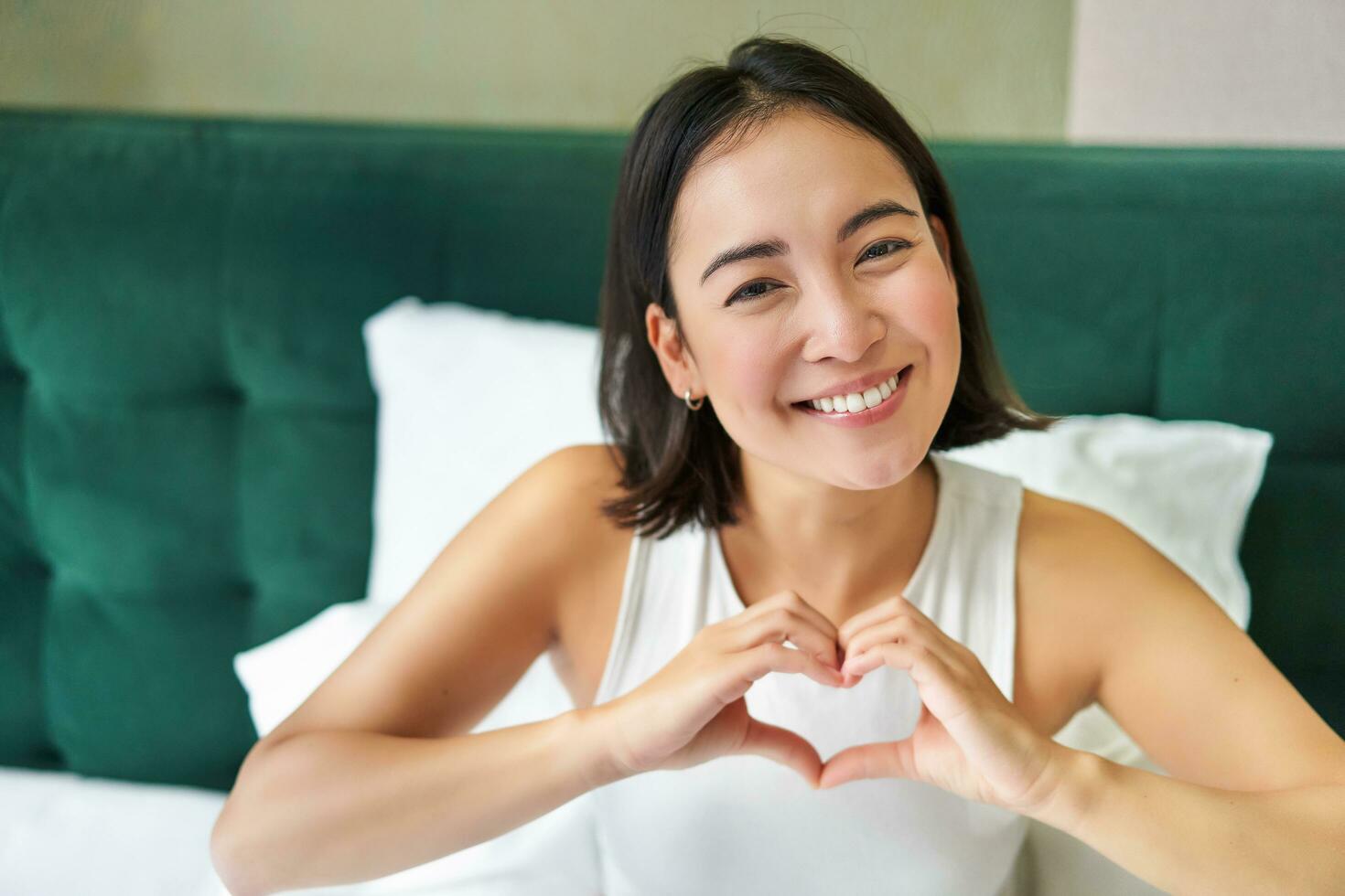 schattig Aziatisch meisje shows hart, liefde teken, aan het liegen in bed en lachend, genieten van ochtenden, lui dagen in slaapkamer foto