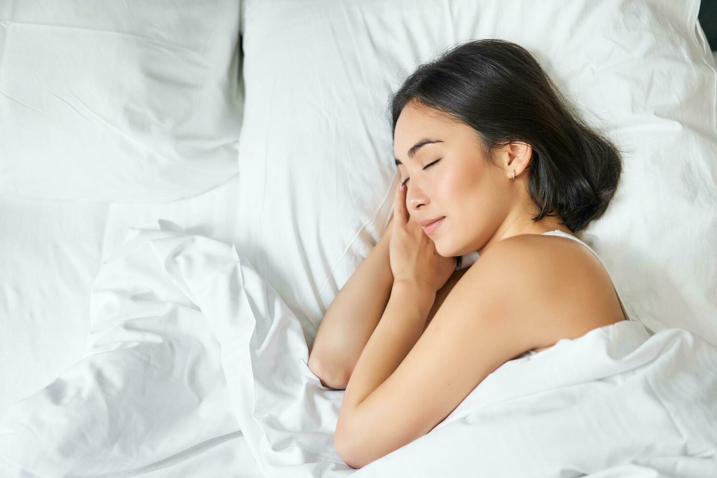top visie beeld van Aziatisch vrouw slapen alleen in koning grootte bed Aan wit kussens. jong meisje aan het liegen in haar slaapkamer met ogen gesloten, ochtend- zonlicht schijnt in kamer foto
