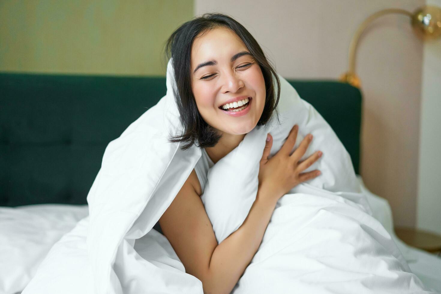 mooi Aziatisch vrouw zittend Aan bed, gedekt met wit donsdeken, lachend, genieten van gelukkig weekend ochtend, lachend Bij camera foto