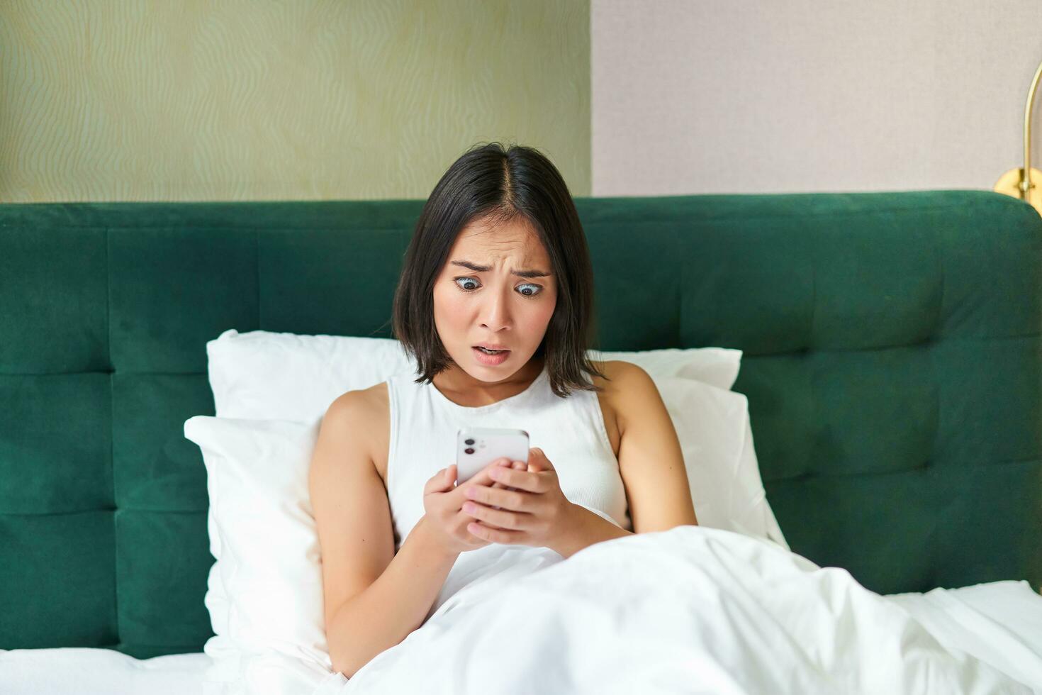 slaapkamer afbeelding van Aziatisch vrouw aan het liegen in bed, op zoek bang en geschokt Bij smartphone scherm, lezing bericht met bezorgd gezicht foto