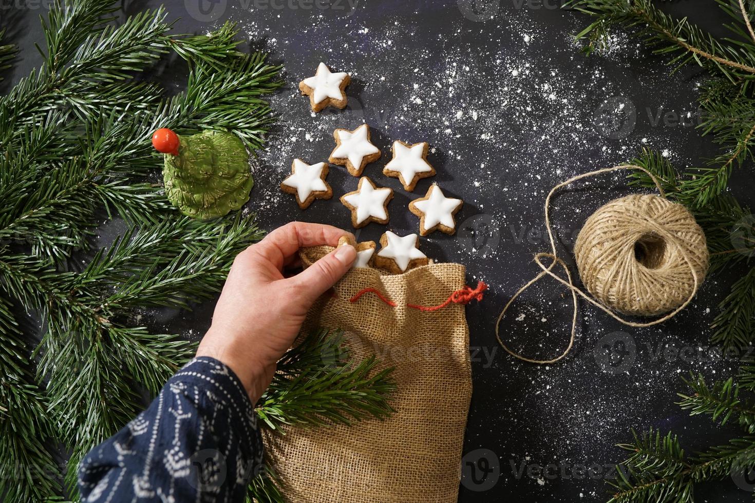 vrouwenhand met traditionele kaneelkoekjes. feestelijke decoratie en kerstboomtakken op de donkere tafel. foto