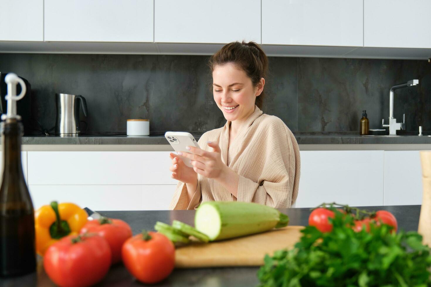 portret van vrouw in badjas zittend in keuken met smartphone, Koken diner, aan het kijken recept Aan sociaal media, video zelfstudie hoe naar bereiden maaltijd foto