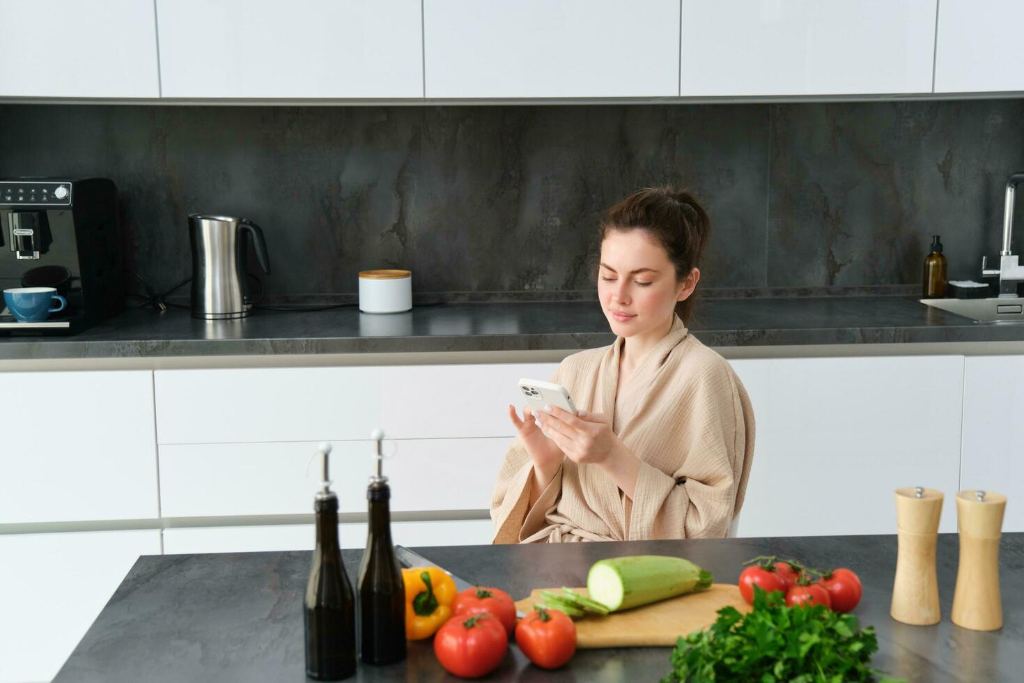 portret van vrouw in badjas zittend in keuken met smartphone, Koken diner, aan het kijken recept Aan sociaal media, video zelfstudie hoe naar bereiden maaltijd foto