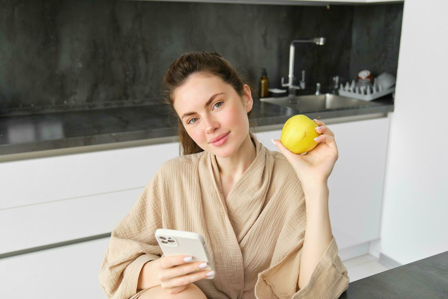 dichtbij omhoog portret van glimlachen brunette vrouw in badjas, zit in keuken Bij huis, toepassingen mobiel telefoon en houdt een appel, bestellingen vers fruit Aan smartphone app, koopt boodschappen online, looks omhoog recept foto