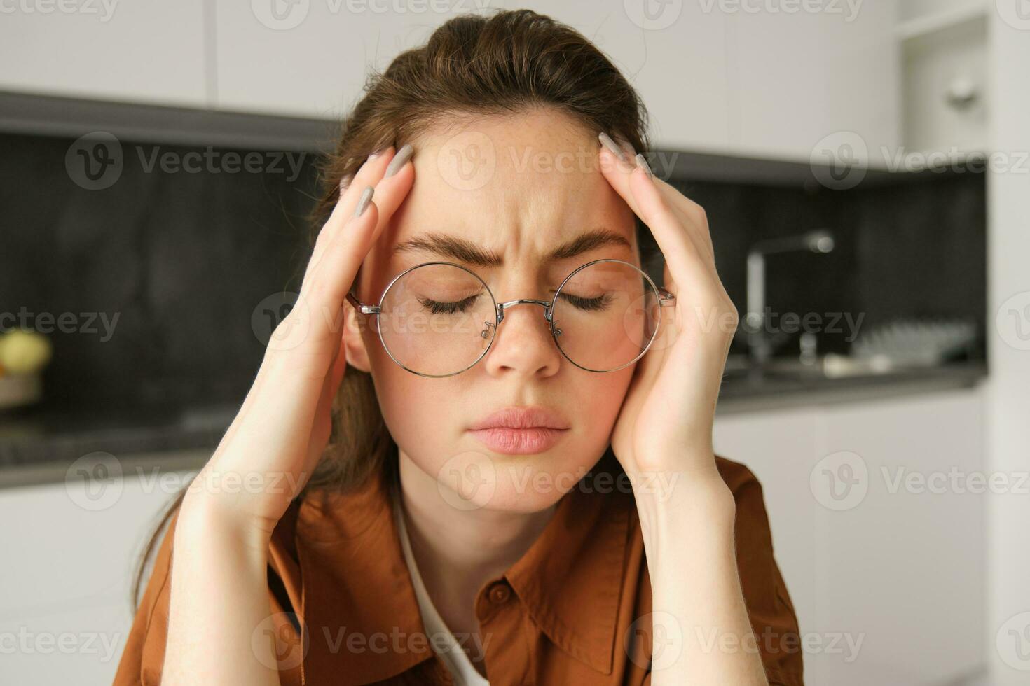 portret van moe jong werken vrouw in bril, houdt handen Aan hoofd, lijdt pijnlijk migraine, heeft pijn in tempels, zittend met hoofdpijn in keuken foto
