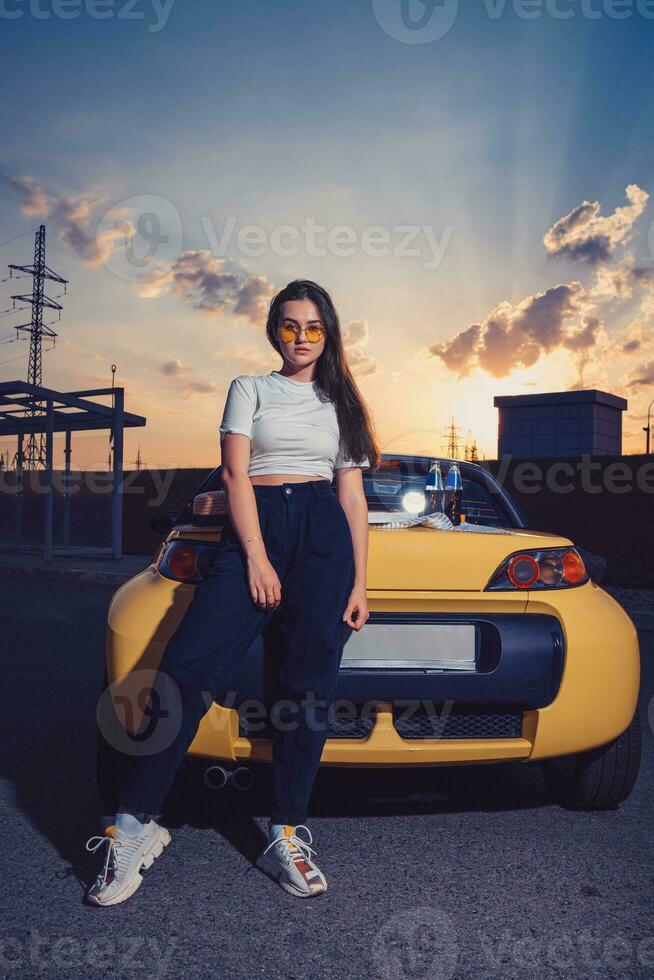 vrouw in zonnebril, wit bovenkant, blauw jeans en sportschoenen is poseren buitenshuis in de buurt geel auto met twee glas flessen van Frisdrank Aan haar kofferbak. bespotten omhoog foto