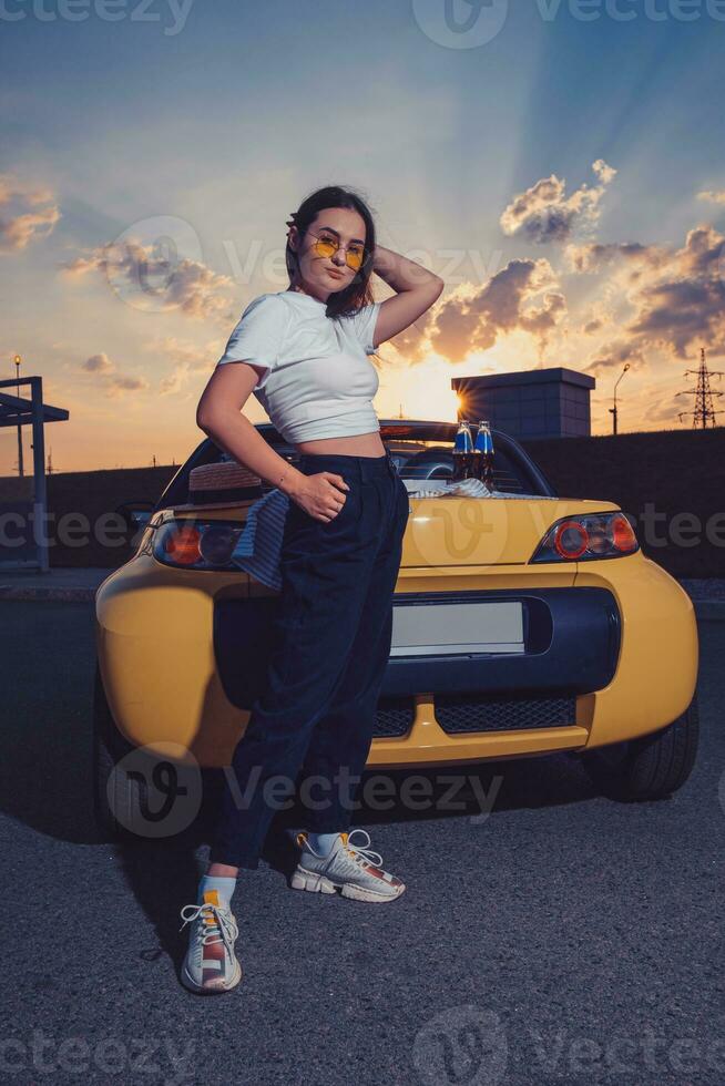 aantrekkelijk meisje in zonnebril, wit bovenkant, zwart broek en sportschoenen is poseren buitenshuis in de buurt geel auto cabriolet met twee flessen van Frisdrank Aan haar romp foto