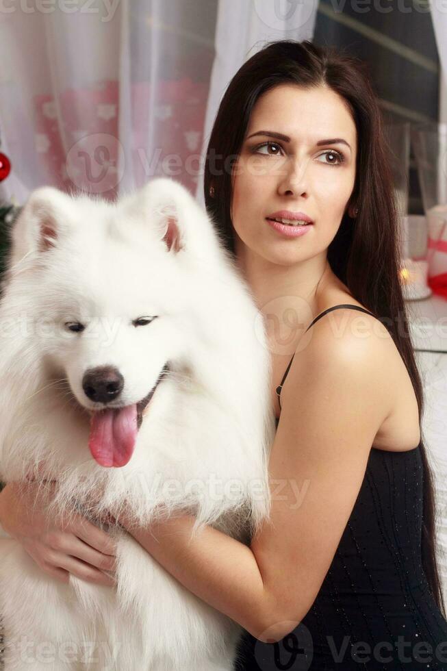 mooi brunette jong vrouw lang zwart jurk met een Sneeuwwitje hond samojeed schor studio in tinten van bruin kaarsen foto