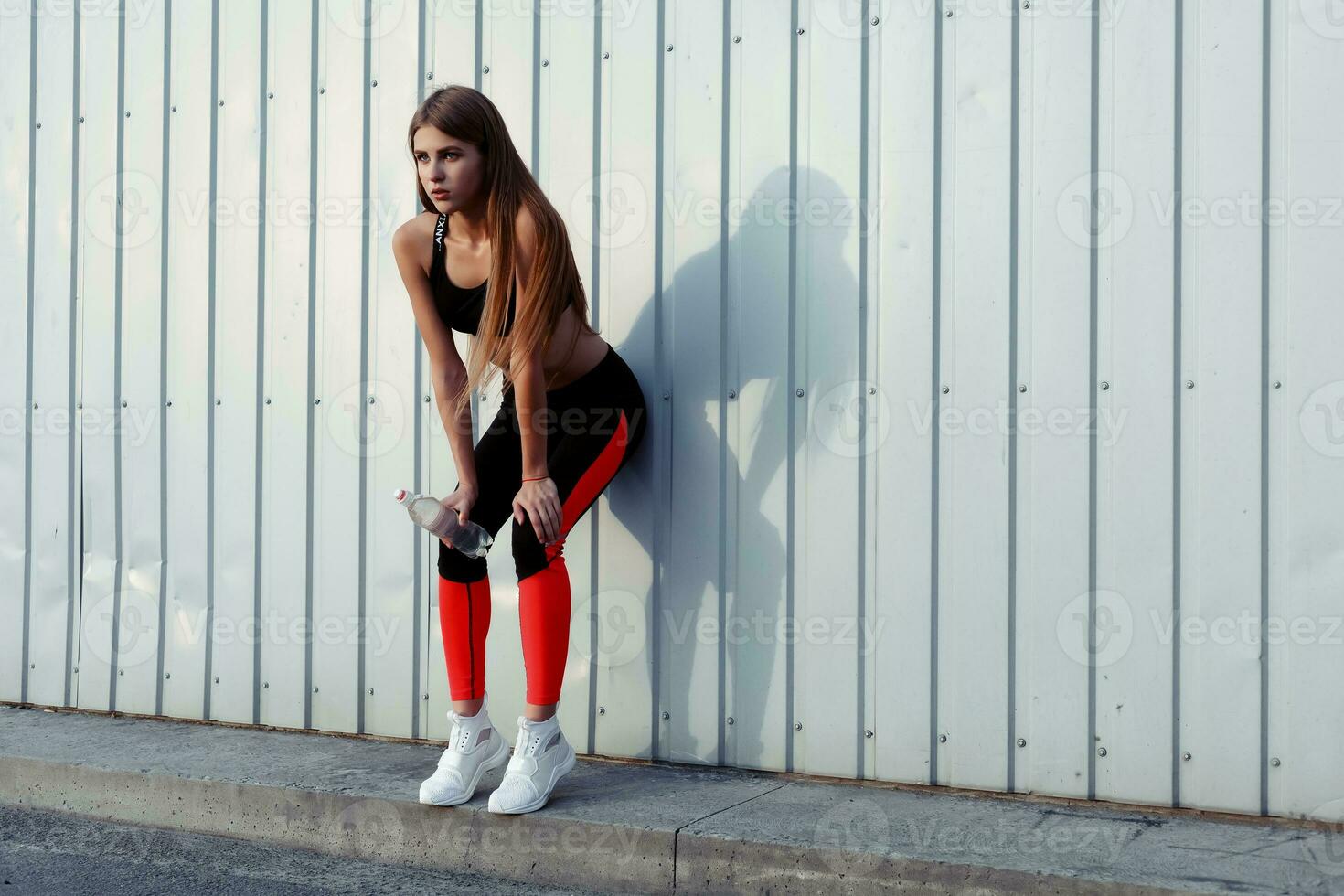vrouw atleet drinken water terwijl staand door een grijs muur. foto