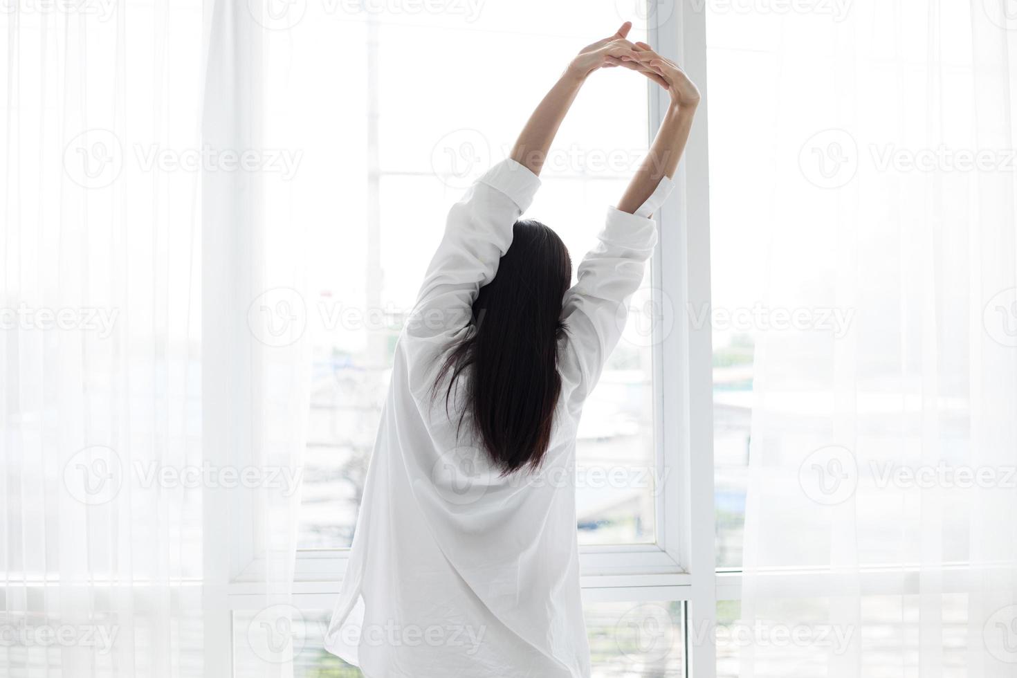 Aziatische vrouw mooie jonge lachende vrouw zittend op bed en strekken zich in de ochtend in de slaapkamer na het wakker worden in haar bed volledig uitgerust en open de gordijnen in de ochtend om frisse lucht te krijgen. foto