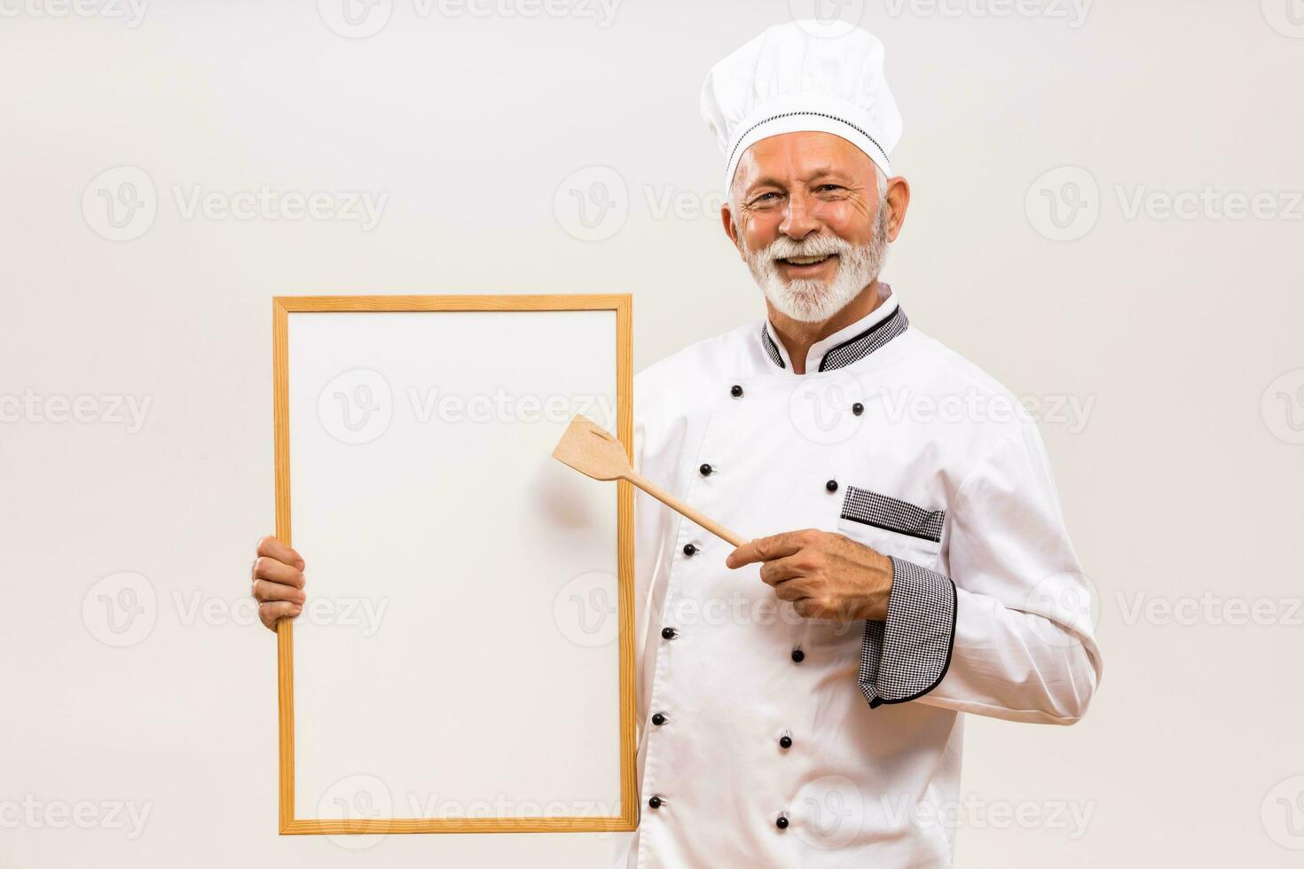 portret van senior chef tonen met houten lepel whiteboard Aan grijs achtergrond. foto