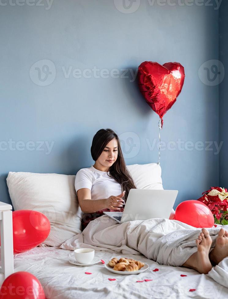 jonge, gelukkige brunette vrouw zit in het bed met rode hartvormige ballonnen die op de laptop werken foto