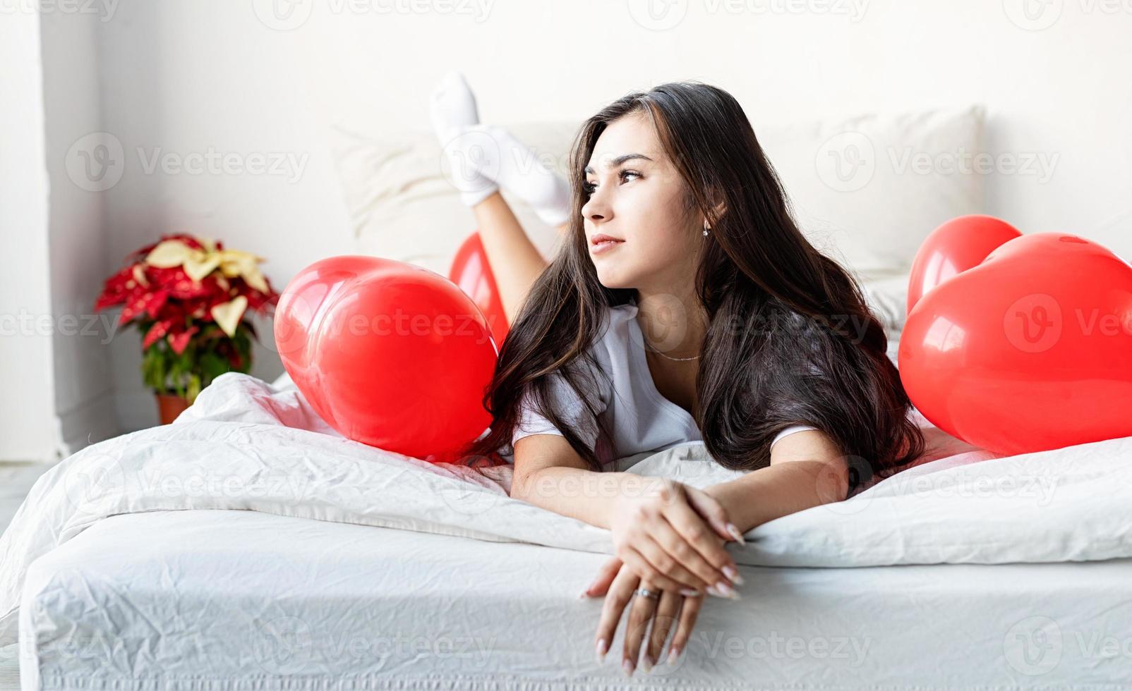 jonge, gelukkige brunette vrouw die in bed ligt met rode hartvormige ballonnen foto