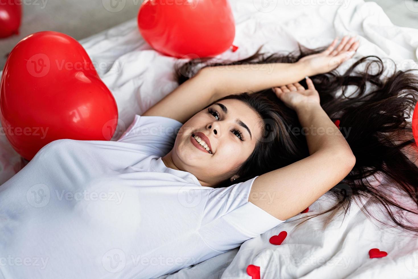 jonge, gelukkige brunette vrouw die in bed ligt met rode hartvormige ballonnen en decoraties foto