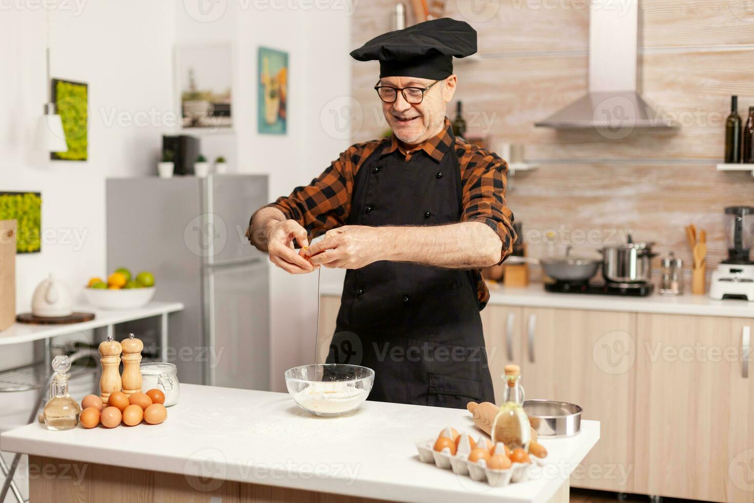 gelukkig senior Mens kraken eieren over- tarwe meel terwijl voorbereidingen treffen traditioneel recept. ouderen gebakje chef kraken ei Aan glas kom voor taart recept in keuken, menging door hand, kneden foto