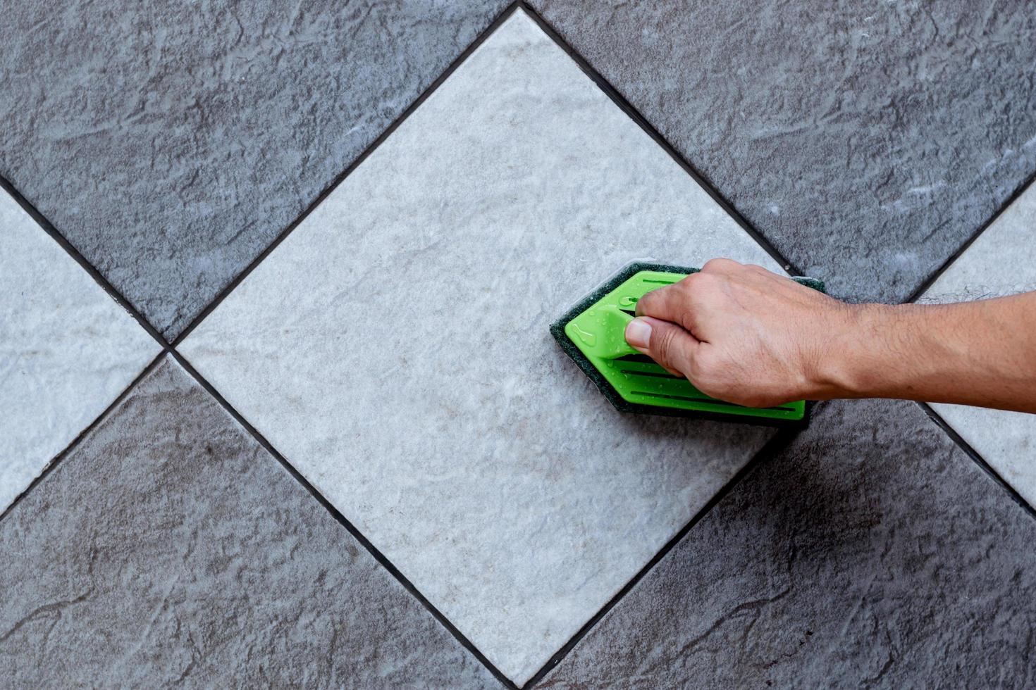 bovenaanzicht van een menselijke hand gebruiken een groene plastic vloerwasser om de tegelvloer te schrobben met een vloerreiniger. foto