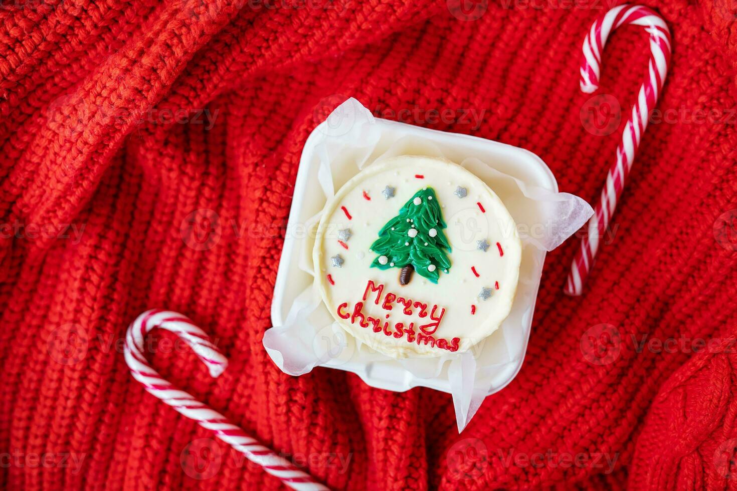 een feestelijk Kerstmis koekje met een wit glimmertjes en een groen boom Aan bovenkant, Aan een rood gebreid achtergrond met snoep stokken. vrolijk Kerstmis belettering. foto