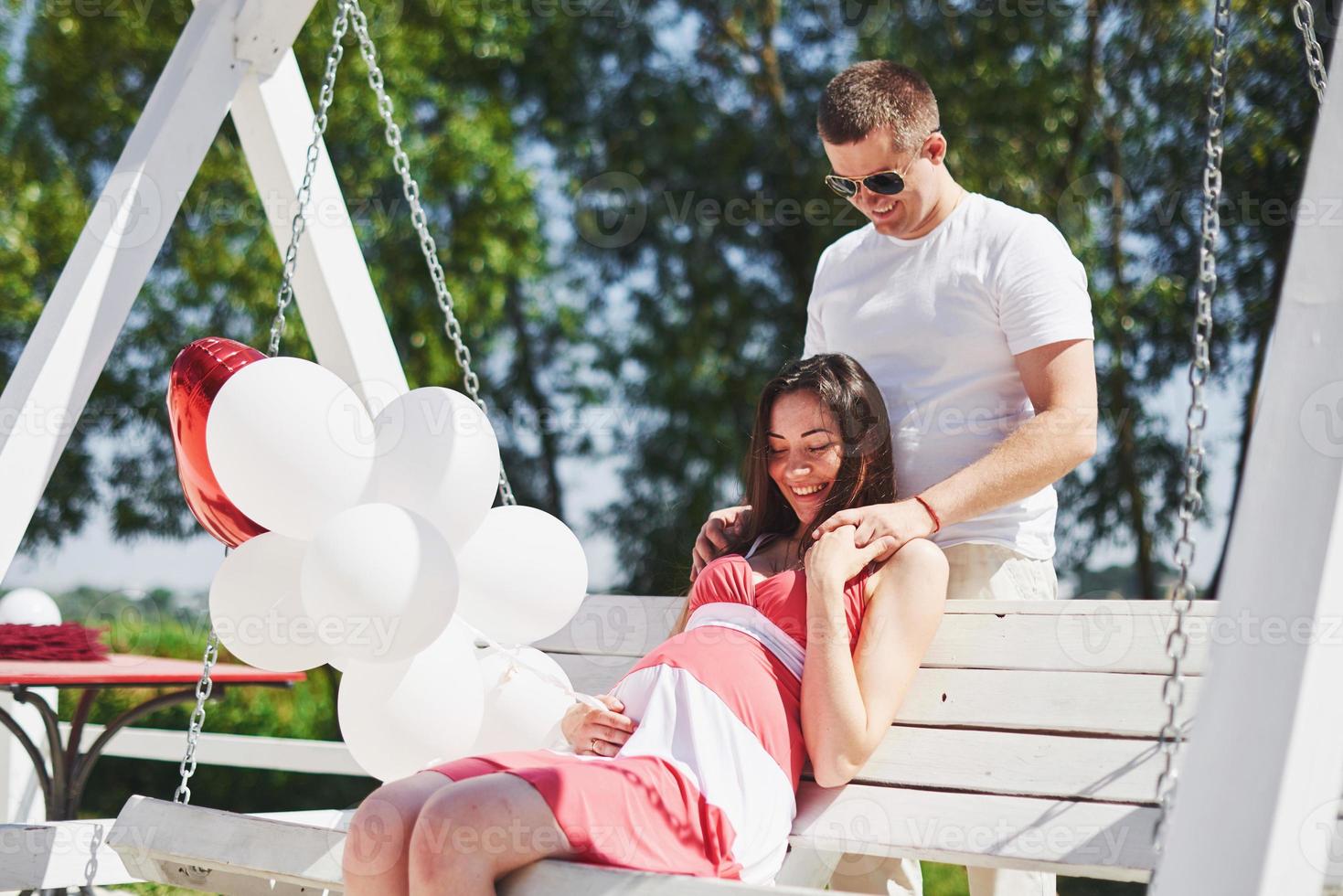 wachten schat. zwangere vrouw met geliefde man zitten op een bankje. man hand omarmt een ronde buik vrouw. ouderschap. zwangere vrouw in een witte jurk in kersen met een rode strik. detailopname. negen maanden foto