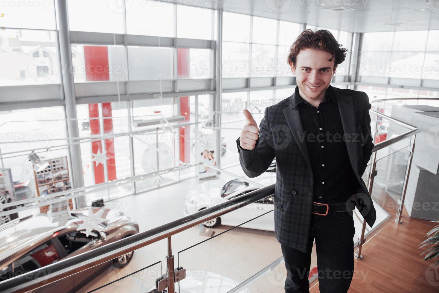 autodealer bezoeken. jonge man op auto salon achtergrond. gelukkig man verschijnt vinger. auto verkoop en verhuur concept foto