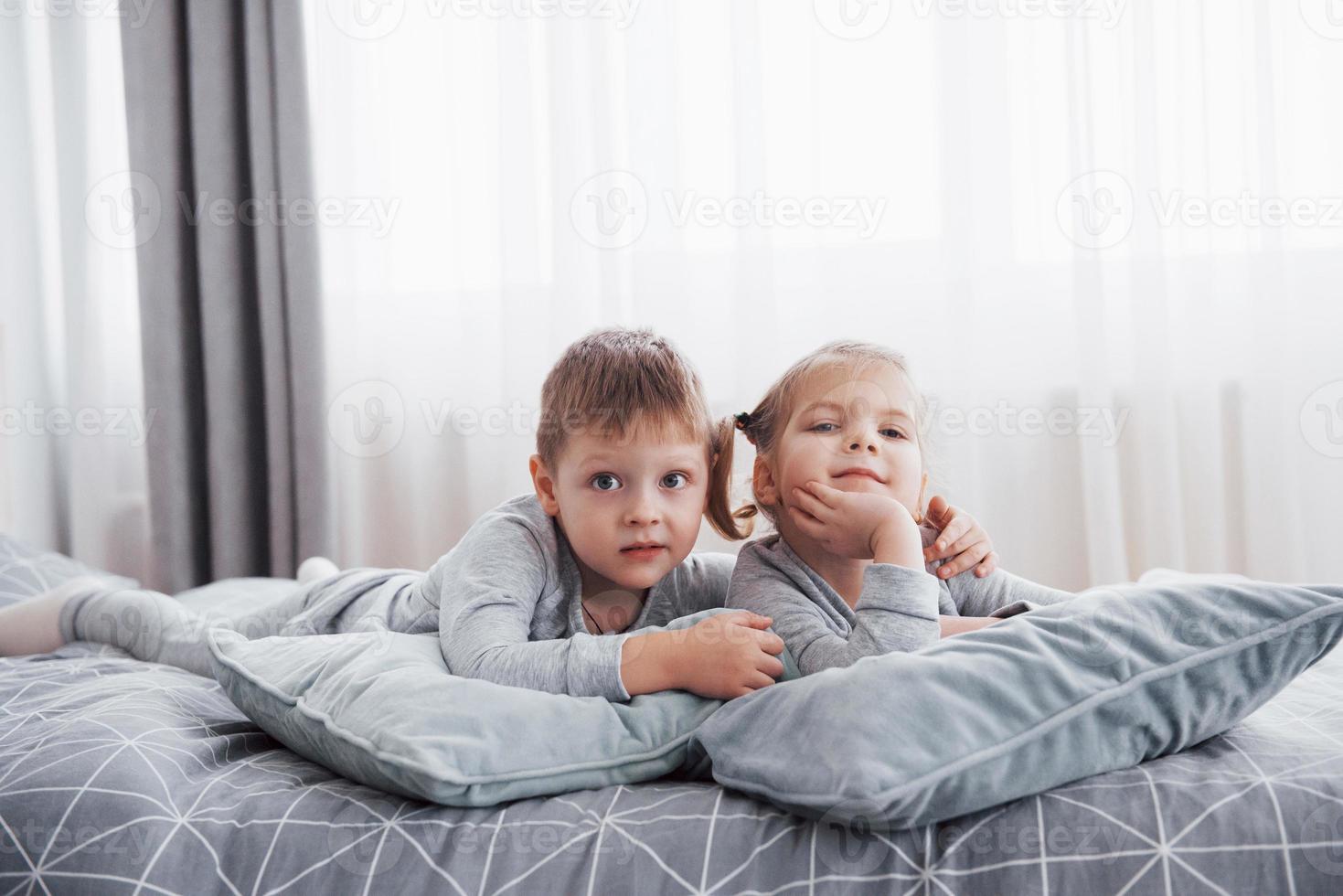 gelukkige jonge geitjes die in witte slaapkamer spelen. kleine jongen en meisje, broer en zus spelen op het bed in een pyjama. kinderkamer interieur voor kinderen. nachtkleding en beddengoed voor baby's en peuters. familie thuis foto