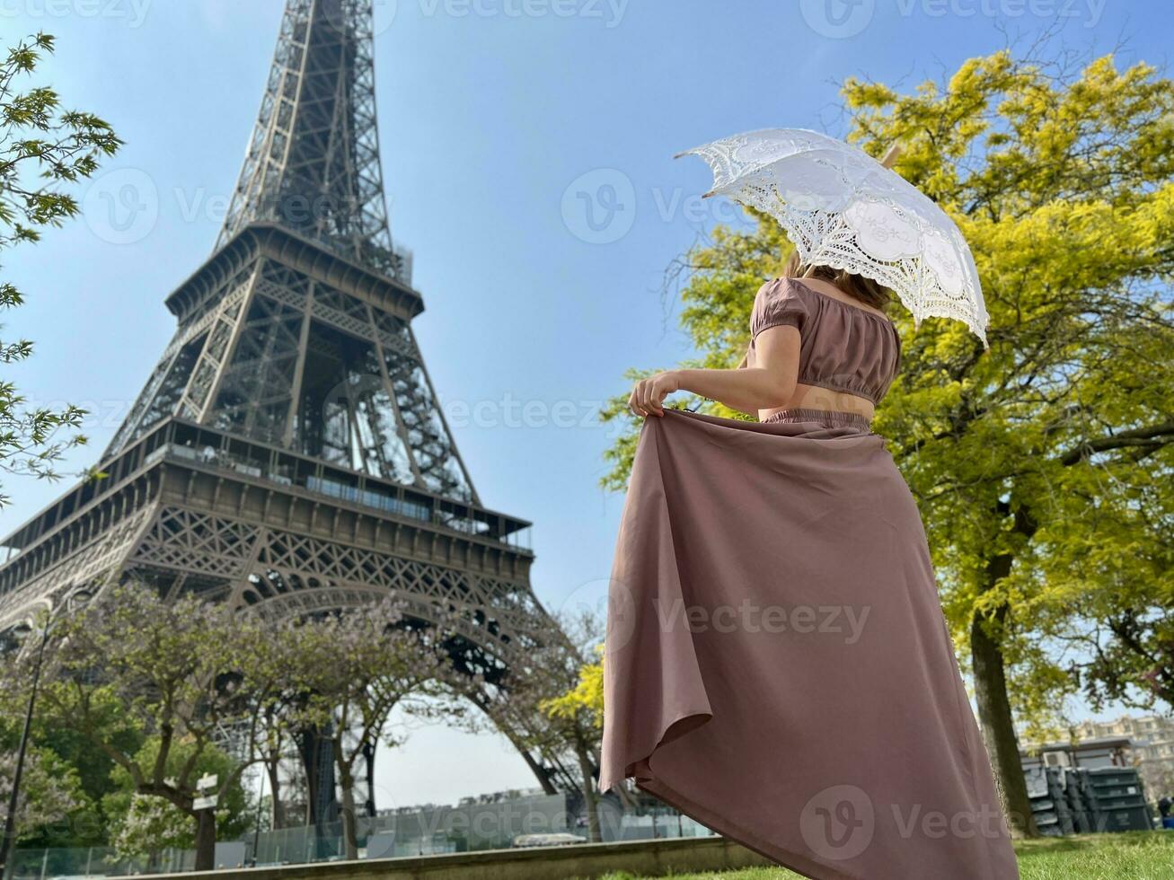 van de terug wij zien een meisje in een mooi lang bruin jurk in een retro stijl met een paraplu wie wandelingen naar de eiffel toren Holding de jurk met een hand- foto