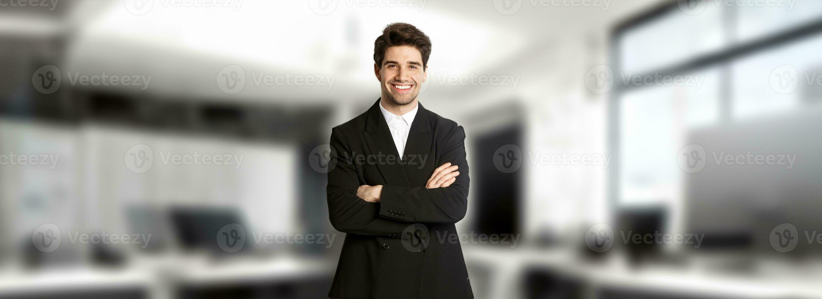 knap en zelfverzekerd gebaard Mens, staand in formeel pak met handen gekruiste Aan borst, glimlachen tevreden met werk, staand over- modern kantoor. foto