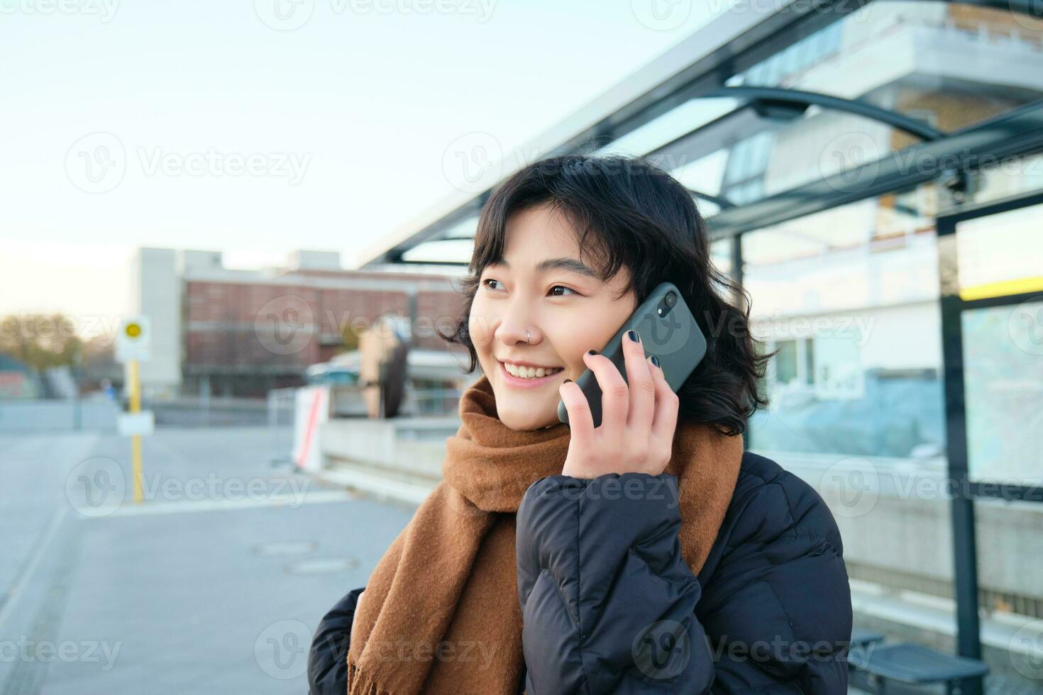 grijnzend Koreaans meisje pratend Aan mobiel telefoon, staand Aan bus hou op, gebruik makend van smartphone, poseren Aan weg in winter, verpakt in sjaal, vervelend zwart jasje foto