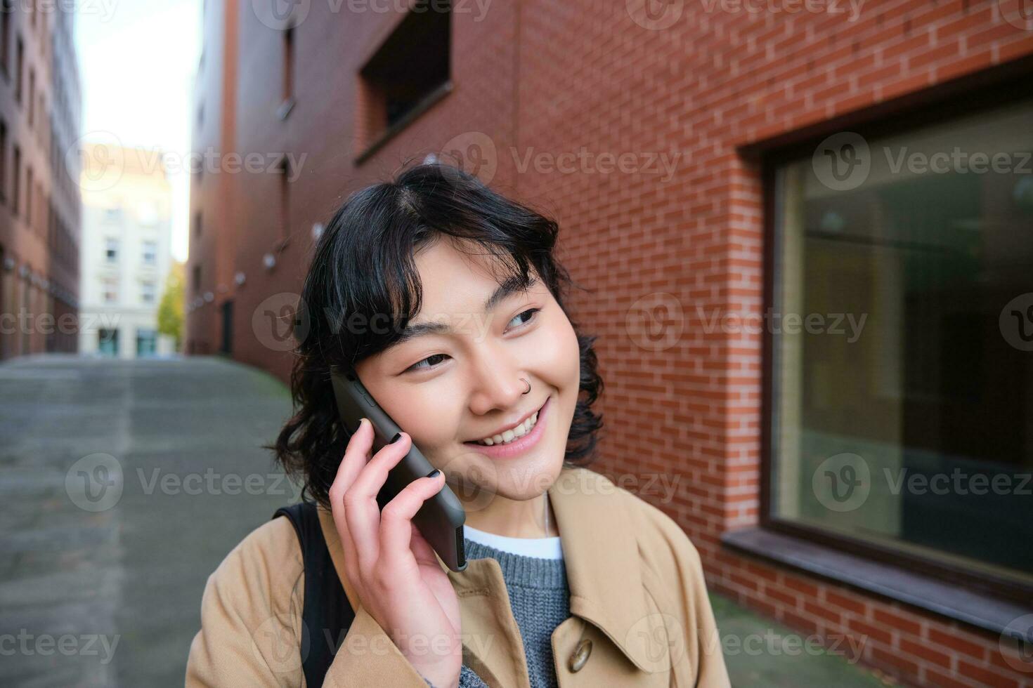 cellulair technologie concept. jong modern meisje praat Aan mobiel telefoon, wandelingen Aan straat en glimlacht, heeft gesprek over- telefoon foto